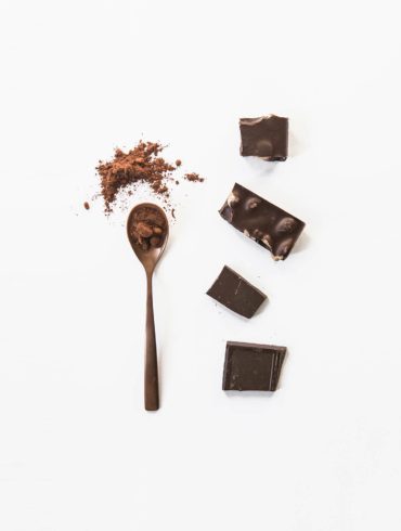 Trozos de chocolate puro con almendras junto a una cuchara con chocolate en polvo sobre fondo blanco. Poppyns Magazine
