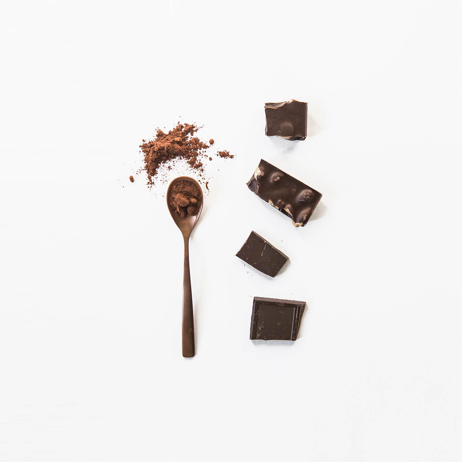 Trozos de chocolate puro con almendras junto a una cuchara con chocolate en polvo sobre fondo blanco. Poppyns Magazine