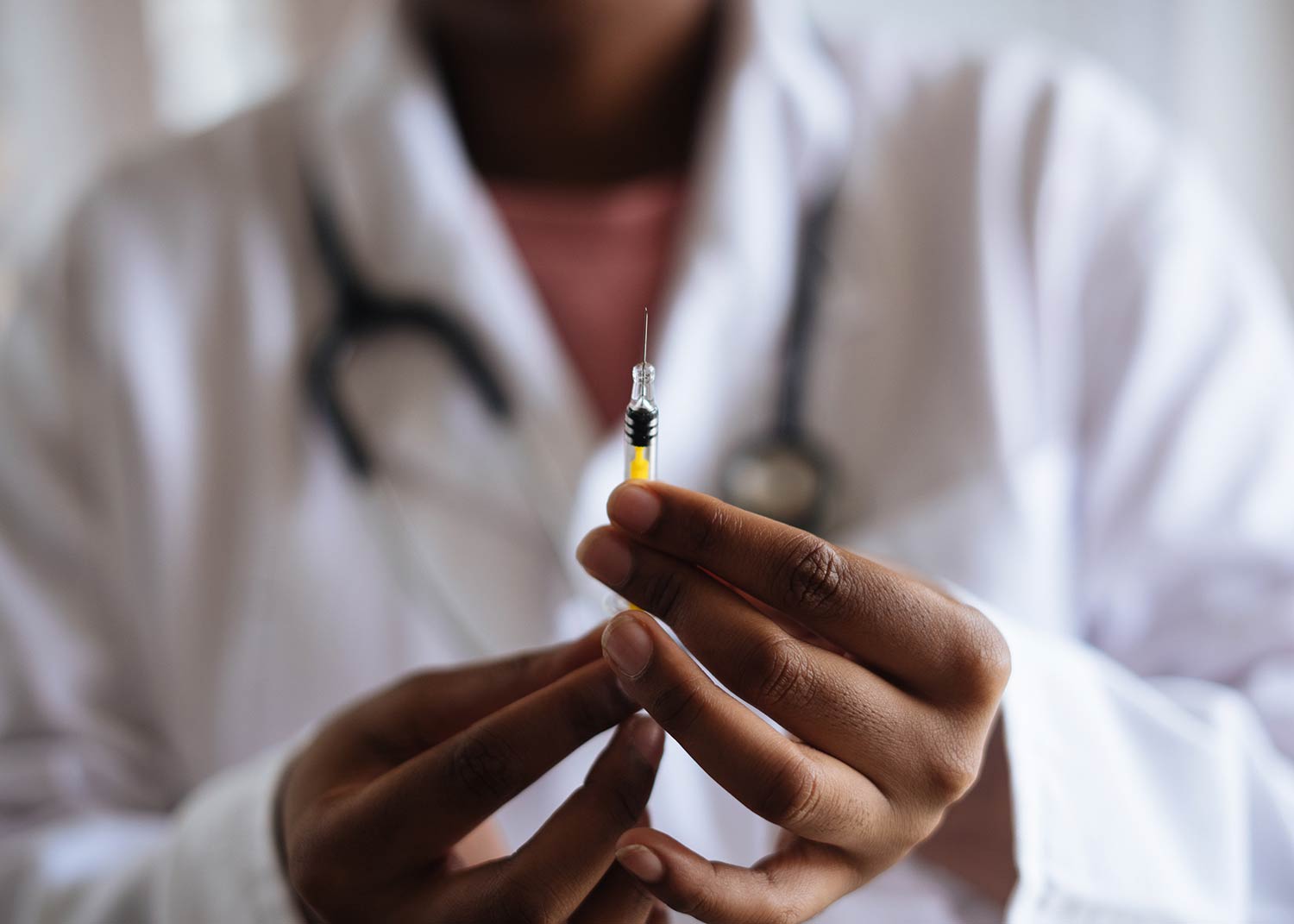 Médico con una bata blanca y un fonendoscopio colgado al cuello, sosteniendo una jeringuilla con una aguja. Vacuna coronavirus. Poppyns Magazine