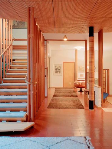 Interior de una vivienda de diseño, con suelo adoquinado color teja, escalera de madera con peldaños enmoquetados, pasamanos de cobre, techo de madera, alfombras y vegetación. Poppyns Magazine
