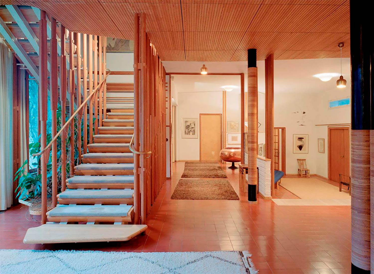 Interior de una vivienda de diseño, con suelo adoquinado color teja, escalera de madera con peldaños enmoquetados, pasamanos de cobre, techo de madera, alfombras y vegetación. Poppyns Magazine