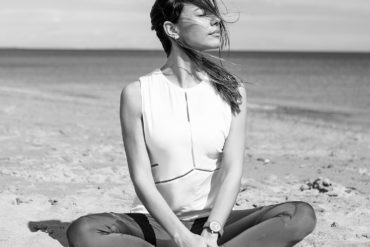 Chica joven morena sentada en la playa sobre la arena, con las piernas cruzadas, los ojos cerrados tomando el sol, el pelo sobre la cara y el mar de fondo.