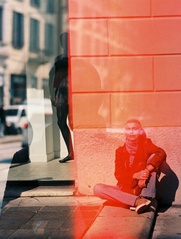 Foto con reflejo en un cristal con un hombre bien vestido sentado en el suelo de la calle con los ojos cerrados y el sol dándole en la cara y la silueta de otro hombre reflejada en el cristal tras una franja roja. Poppyns Magazine