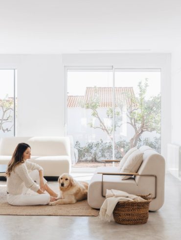 Salón minimalista con muebles de diseño, un cesto de mimbre con mantas, una mujer sentada sobre una alfombra junto a un perro golden blanco y un ventanal con un jardín al fondo. Poppyns Magazine