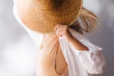 Mujer rubia con un bañador de rayas, pantalón de lino beige y camisa de lino blanca, tapándose el rostro con un sombrero de mimbre. Poppyns Magazine.