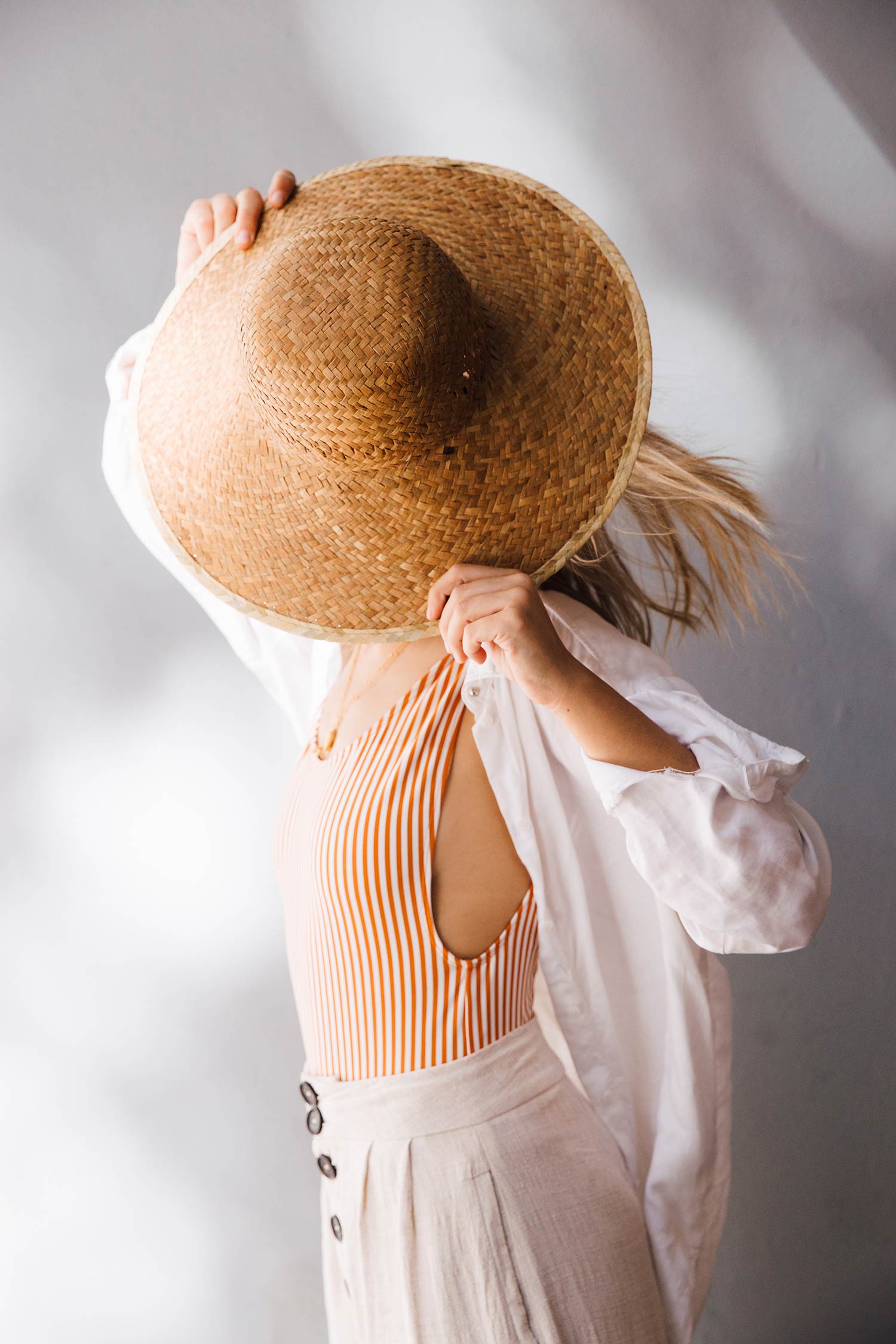 Mujer rubia con un bañador de rayas, pantalón de lino beige y camisa de lino blanca, tapándose el rostro con un sombrero de mimbre. Poppyns Magazine.