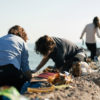 Personas agachadas recogiendo residuos en la playa entre las rocas. Poppyns Magazine