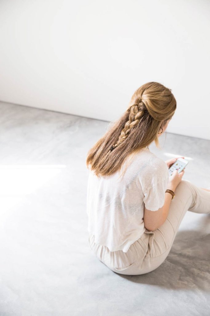 Chica sentada de espaldas mirando el móvil en una habitación de diseño minimalista con suelo de hormigón en época de cuarentena. Poppyns Magazine