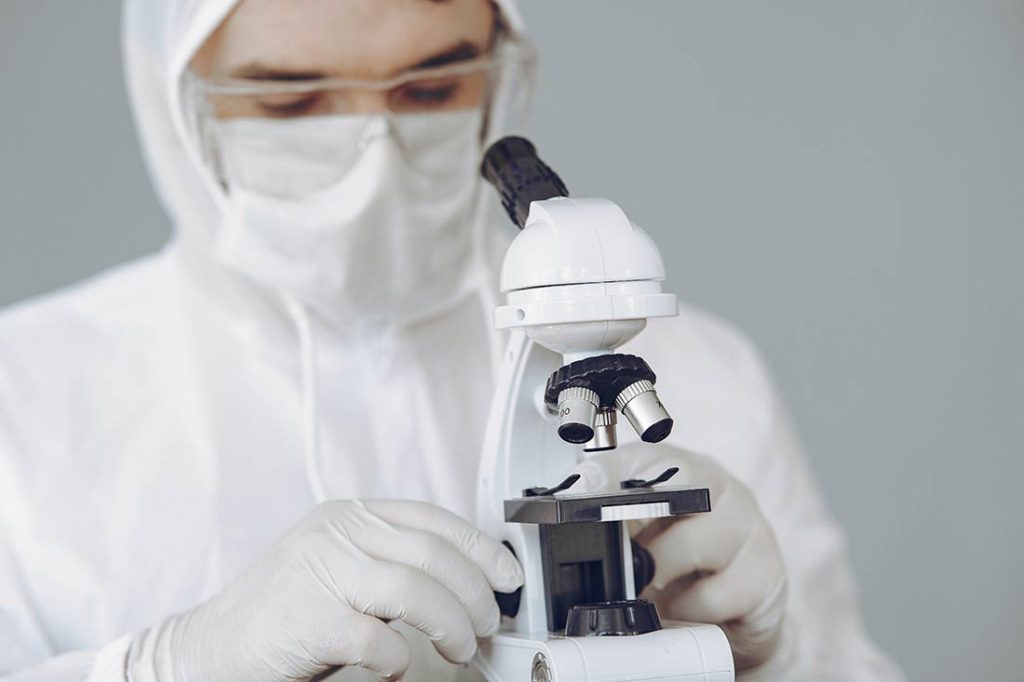 Investigador con gafas de laboratorio, mascarilla, guantes y escafandra blanca, mirando por un microscopio. Poppyns Magazine