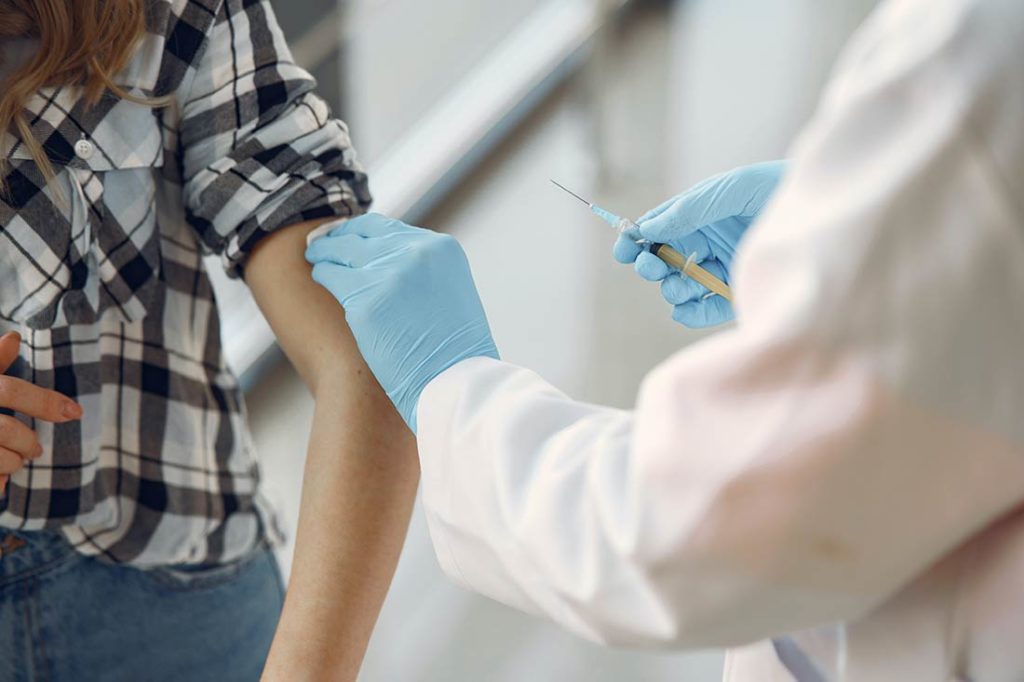 Persona siendo vacunada por un doctor con bata blanca y guantes de latex azules sosteniendo una jeringa. Poppyns Magazine