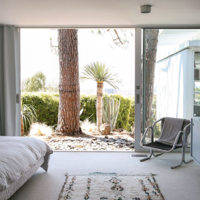Dormitorio decorado estilo nórdico con muebles de diseño y un ventanal corredero que da a un jardín con piedras y vegetación. Poppyns Magazine
