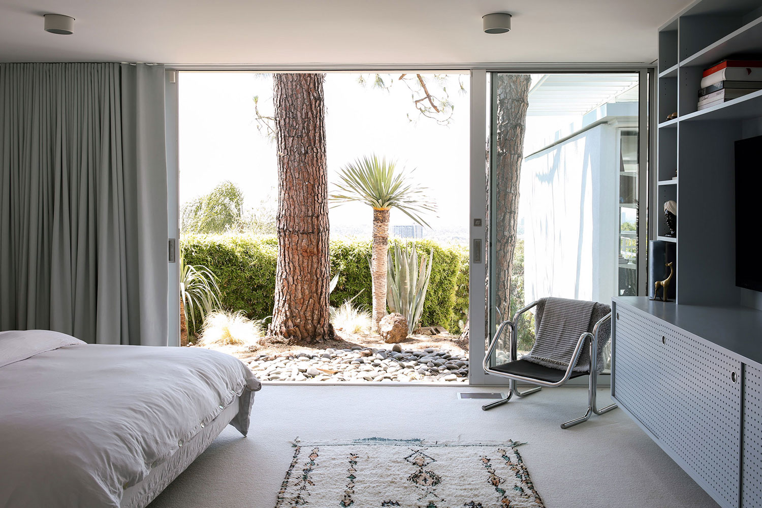 Dormitorio decorado estilo nórdico con muebles de diseño y un ventanal corredero que da a un jardín con piedras y vegetación. Poppyns Magazine
