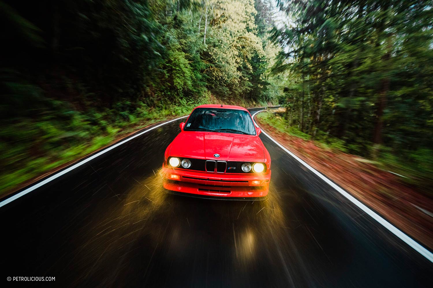BMW M3 rojo circulando lloviendo por una carretera de montaña entre árboles. Poppyns Magazine