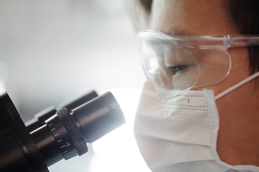 Persona mirando a través de un microscopio con mascarilla y gafas de laboratorio. Poppyns Magazine