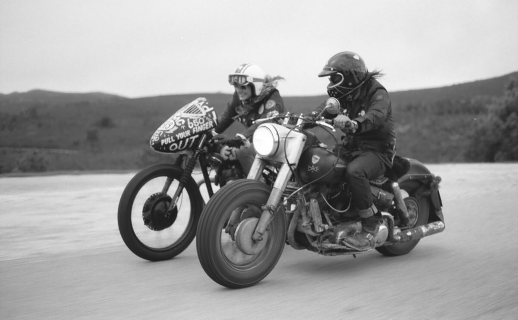 Foto en blanco y negro de dos moteros con motos customizadas rodando por una carretera de tierra con montañas al fondo. Poppyns Magazine