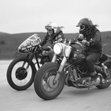 Foto en blanco y negro de dos moteros con motos customizadas rodando por una carretera de tierra con montañas al fondo. Poppyns Magazine