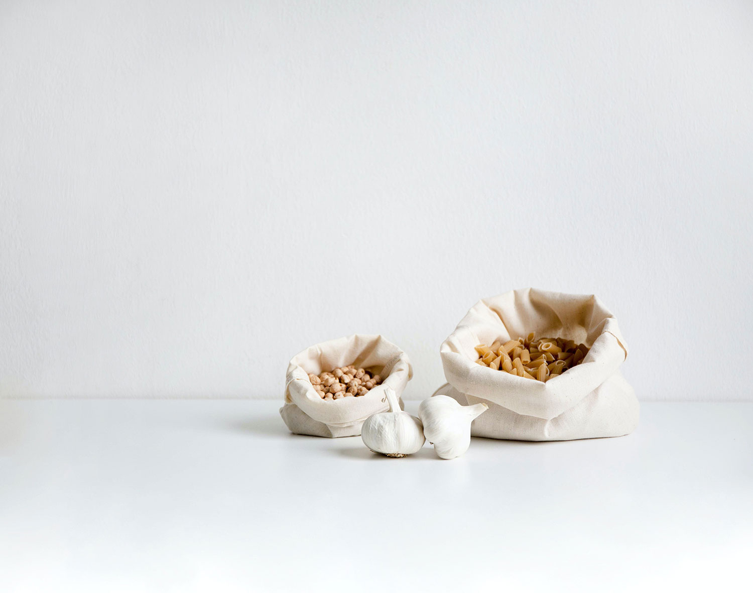 Bolsas de tela blanca con pasta y garbanzos junto con dos ajos. Poppyns Magazine