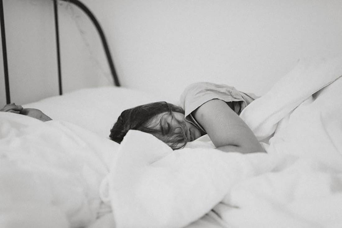 Foto en blanco y negro de mujer durmiendo en la cama de lado tapada con el edredón. Poppyns Magazine