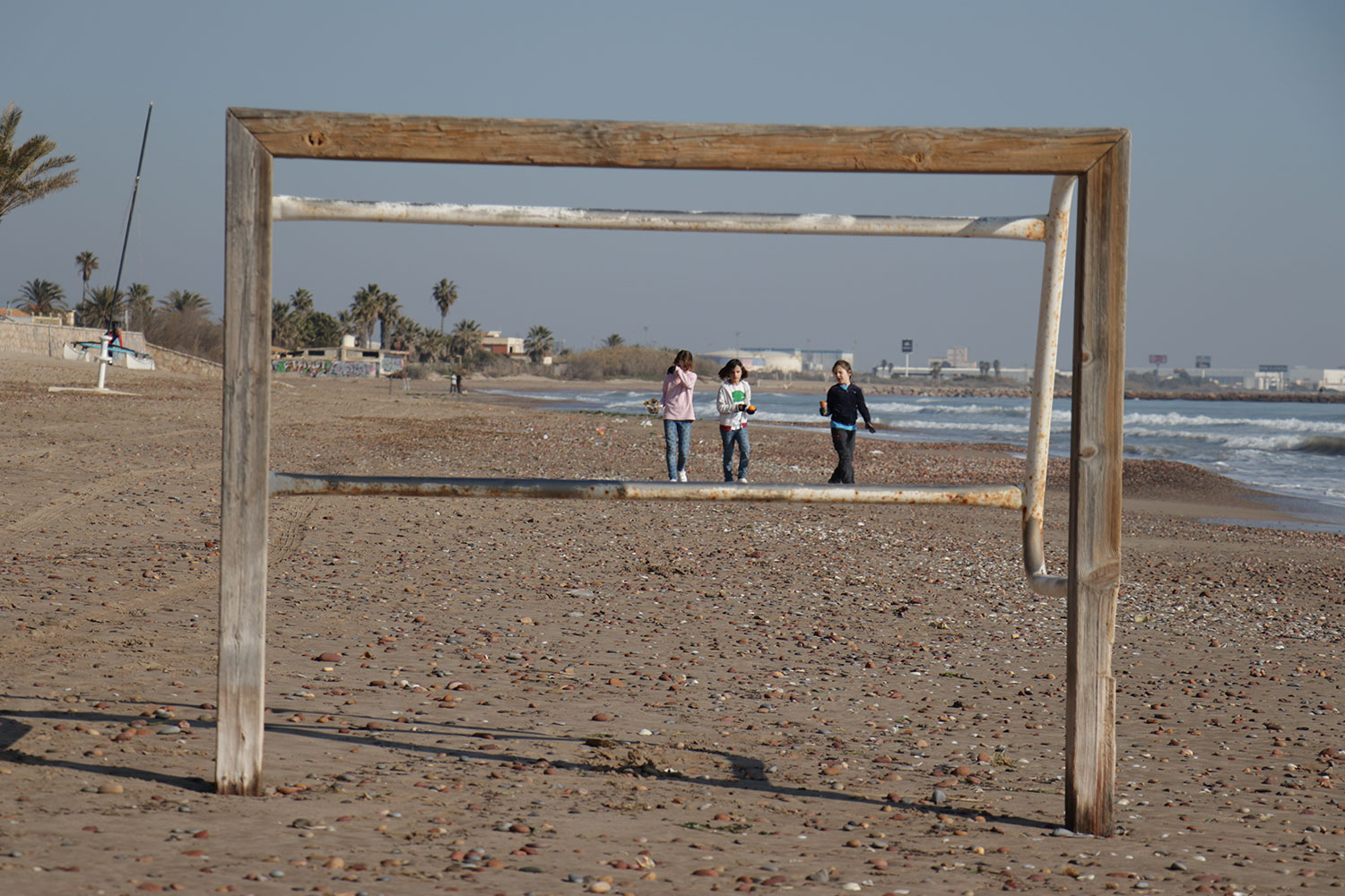 Portería de fútbol de madera en la playa con tres niñas paseando detrás. Poppyns Magazine