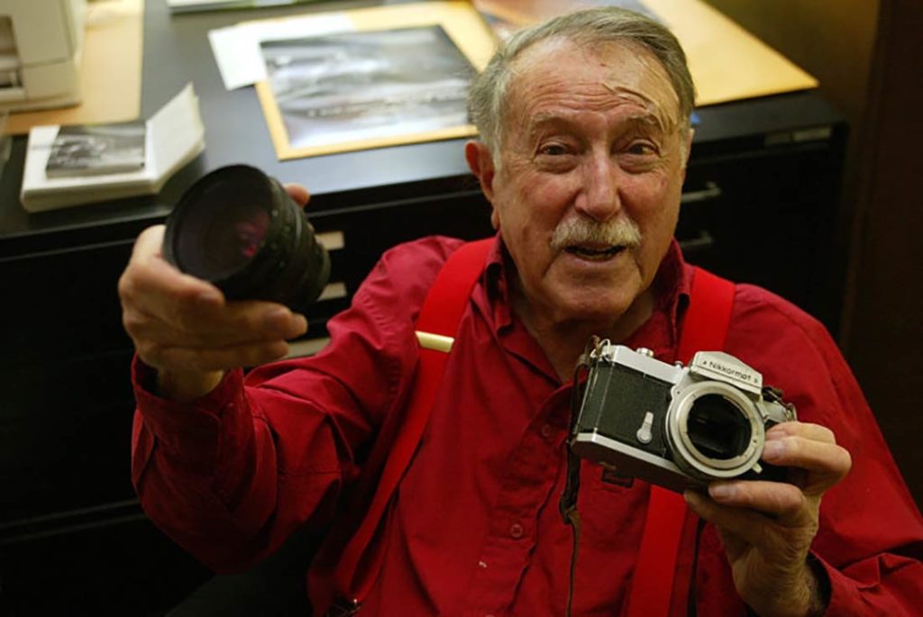 Señor mayor con bigote, vistiendo una camisa roja con tirantes rojos, sonriendo mostrando una cámara de fotos y su objetivo. Poppyns Magazine