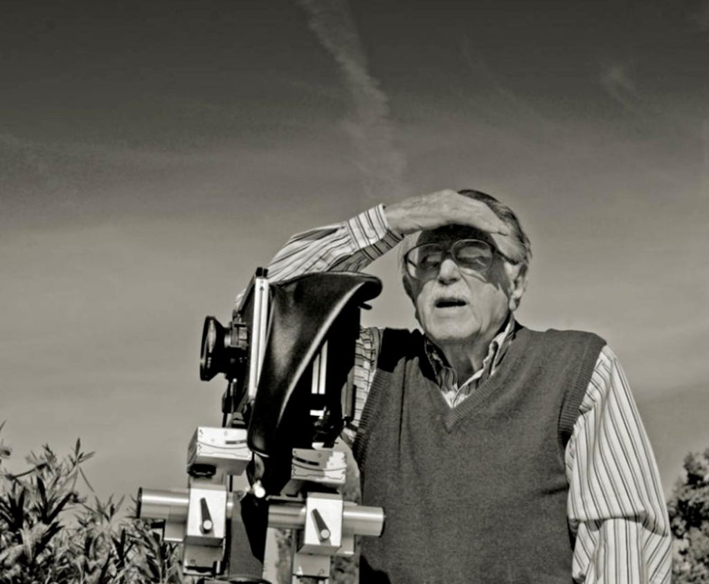 Foto en blanco y negro de un señor mayor con gafas, bigote, camisa a rayas y chaleco, mirando al horizonte tapándose el sol con la mano, junto a una cámara de fotos. Poppyns Magazine