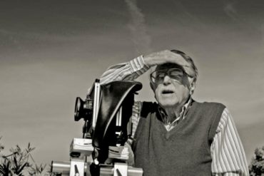 Foto en blanco y negro de un señor mayor con gafas, bigote, camisa a rayas y chaleco, mirando al horizonte tapándose el sol con la mano, junto a una cámara de fotos. Poppyns Magazine