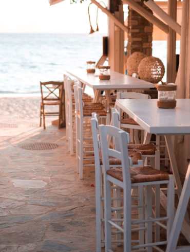 Terraza con sillas altas de madera blanca y mimbre en un restaurante a pie de mar en una calle empedrada. Poppyns Magazine
