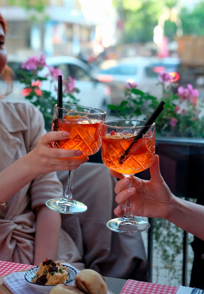 Dos personas brindado con una copa en la mano en una terraza. Poppyns Magazine