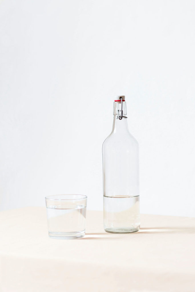 Botella de agua de cristal con tapón y vaso de cristal lleno de agua. Poppyns Magazine