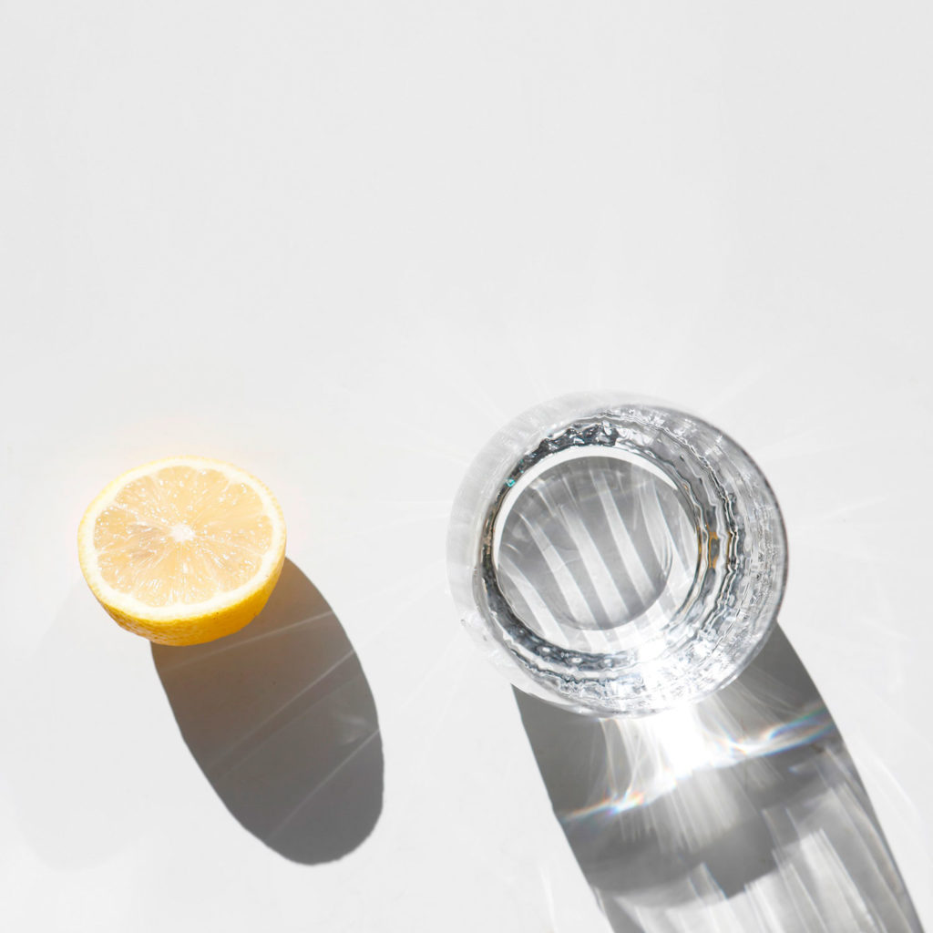 Vaso de de cristal lleno de agua junto con un limón y sus sombras proyectadas. Poppyns Magazine