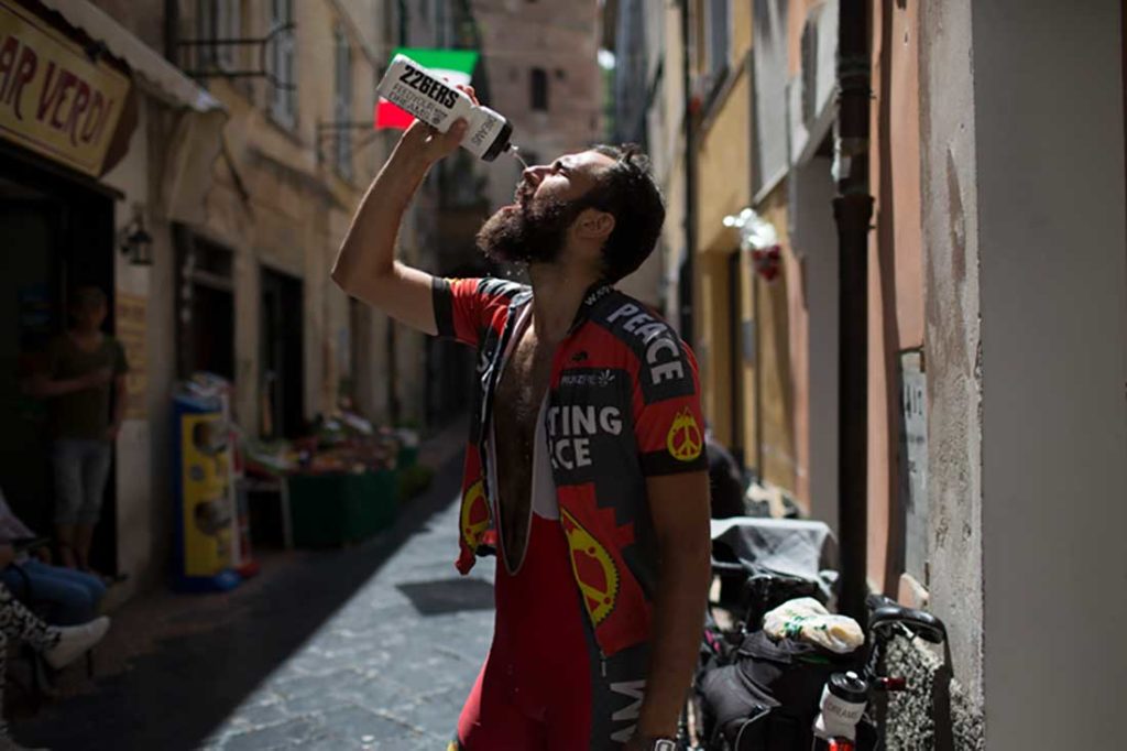 Ciclista con el mallot abierto mojándose la cara con una botella en una calle empedrada. Poppyns Magazine