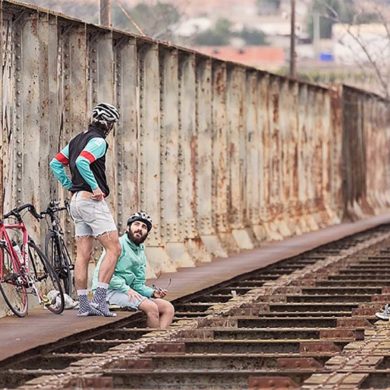 Dos ciclistas hablando sentados en la vía de un tren con las bicis apoyadas en una estructura metálica. Poppyns Magazine