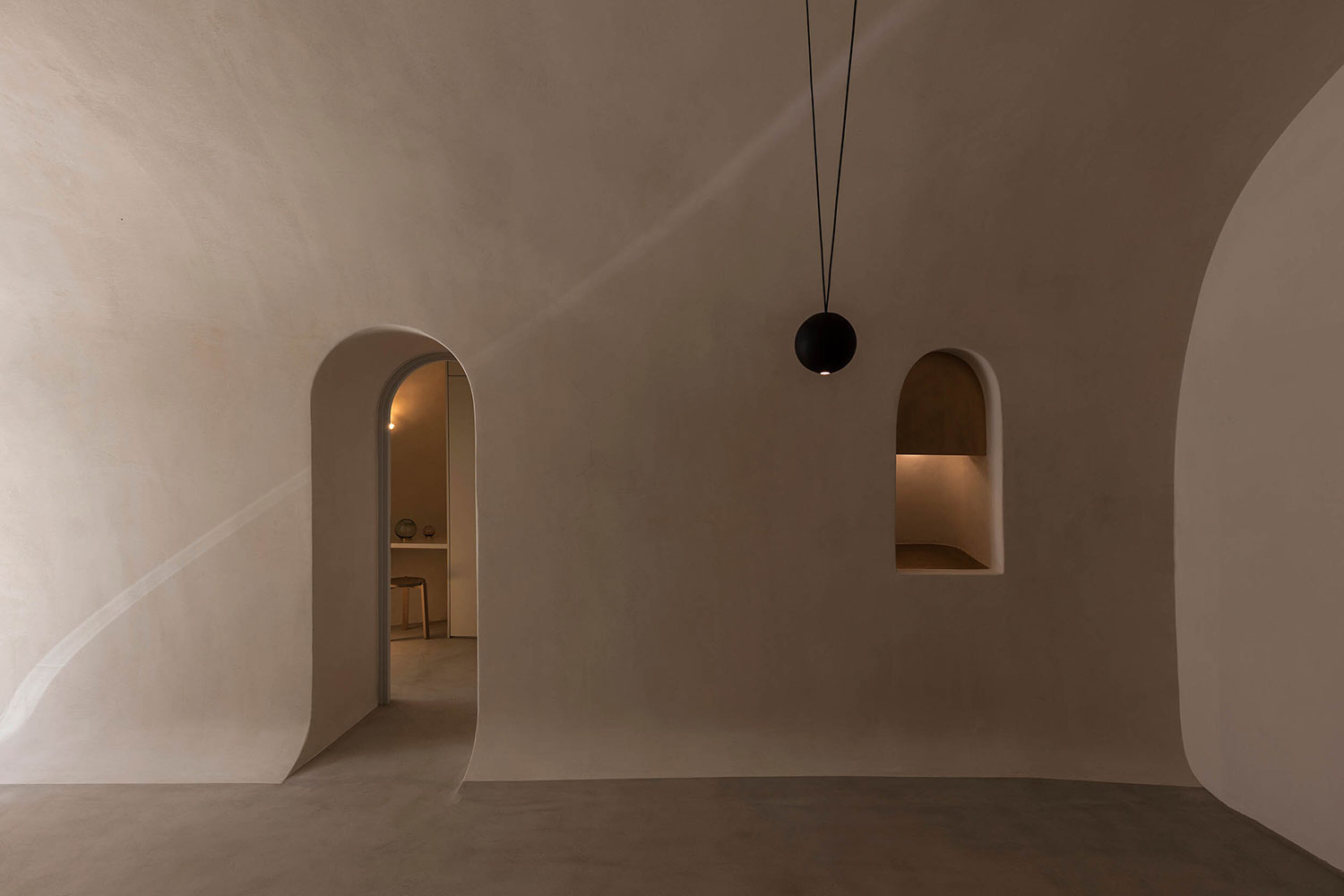 Interior de vivienda minimalista con paredes lisas en tono crema y suelo de microcemento, con una ventana y una puerta con arco ovalado. Poppyns Magazine