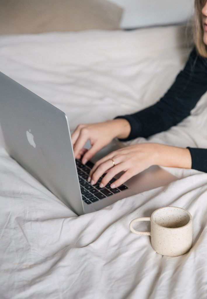 Mujer escribiendo en un ordenador Mac plateado encima de una cama con una taza de te al lado. Poppyns Magazine