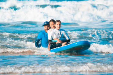 Tres personas, un chico con gorra, una chica y una niña con una camiseta que pone "kind surf", sentadas encima de una tabla de surf dentro del mar. Poppyns Magazine