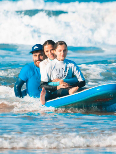Tres personas, un chico con gorra, una chica y una niña con una camiseta que pone "kind surf", sentadas encima de una tabla de surf dentro del mar. Poppyns Magazine