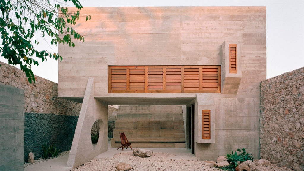 Edificio arquitectónico de hormigón y madera con formas geométricas, ventanales de madera con mallorquinas y zona ajardinada. Poppyns Magazine