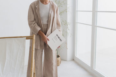 Persona sujetando una revista con portada blanca y con texto 'New York Rises', vestida con pantalón ancho, camiseta blanca y chaqueta tres cuartos en una habitación blanca con un ventanal. Poppyns Magazine