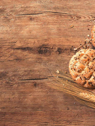 Hogaza de pan con semillas de trigo y sésamo junto a espigas de trigo sobre una mesa de madera. Poppyns Magazine