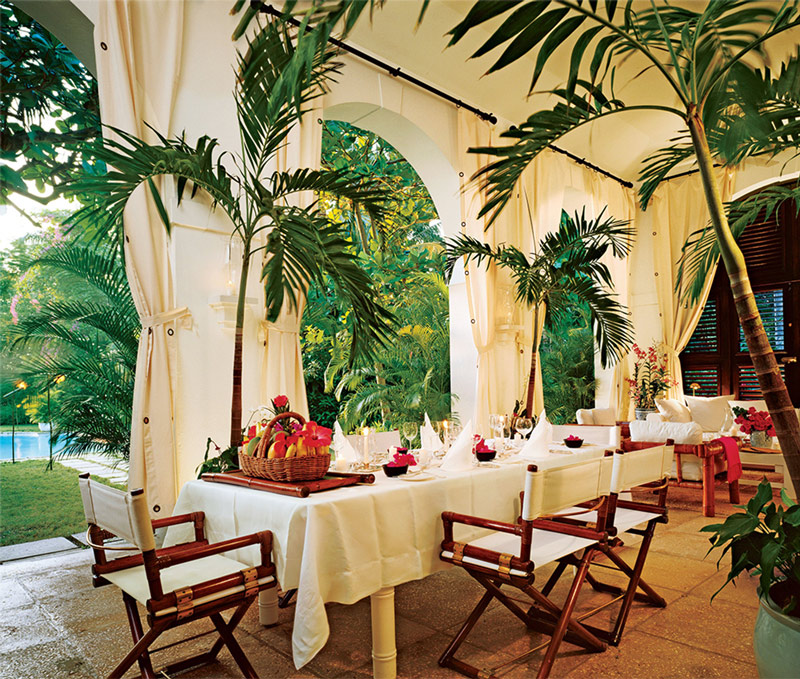 Terraza mansión con mesa para comer frente a jardín con piscina en Jamaica.