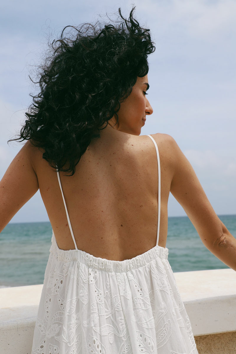 Mujer morena de espaldas frente al mar con vestido blanco estilo mediterráneo.